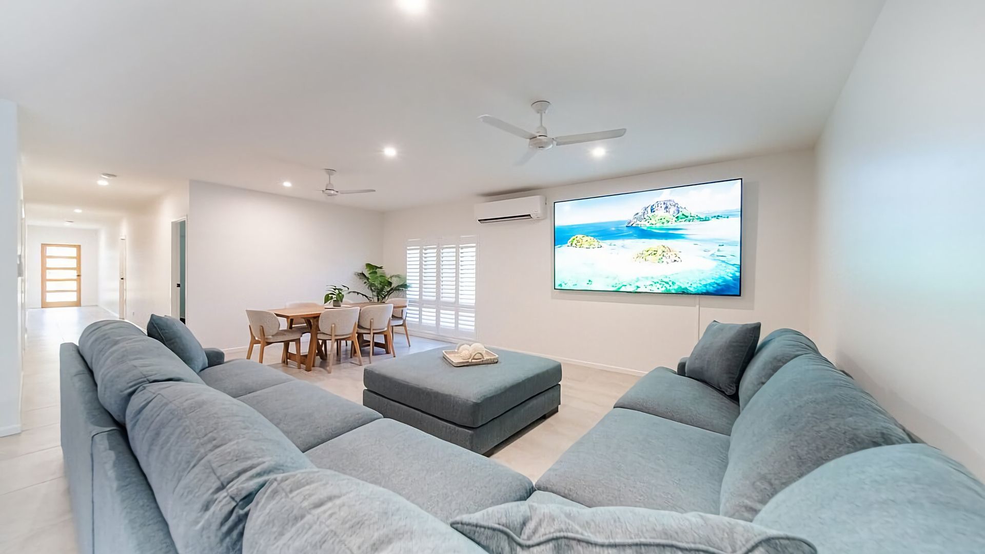 Een moderne woonkamer met een grijze modulaire bank, een eethoek met een houten tafel en stoelen, en een grote flatscreen televisie aan de muur.