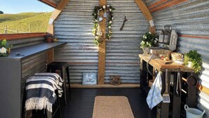 A rustic interior with a corrugated metal wall, wooden beams, a bench, a table with kitchen utensils, and a view of the countryside. - Field and Fleece Farm Stay by Tiny Away (Levels Valley)