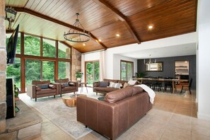 A spacious living room with leather sofas, a dining area, and large windows offering a view of greenery. - Luxury Smoky Mountain Retreat | Theater, Firepit, Hot Tub & Outdoor Movies (Pigeon Forge)