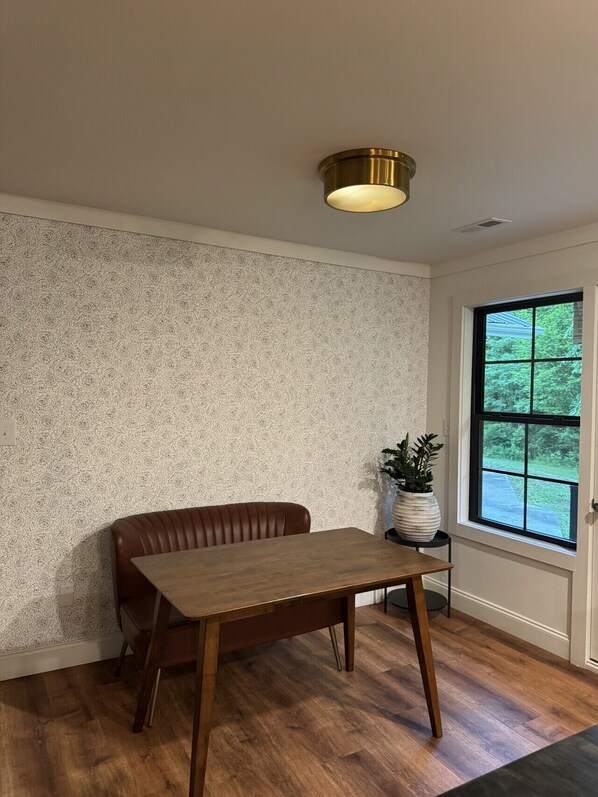 A room with a patterned wallpaper, a wooden table, a brown leather bench, a potted plant, and a window with a view of greenery. - Luxury Smoky Mountain Retreat | Theater, Firepit, Hot Tub & Outdoor Movies (Pigeon Forge)