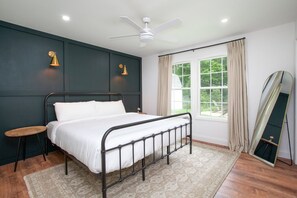 Un dormitorio con cama, un taburete pequeño, un ventilador de techo, una ventana con cortinas y un espejo.