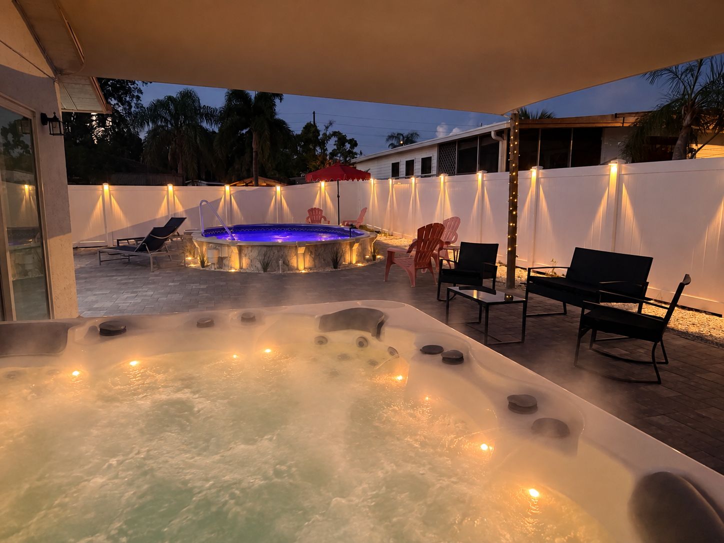A hot tub area with seating and a pool, surrounded by a white fence and palm trees.