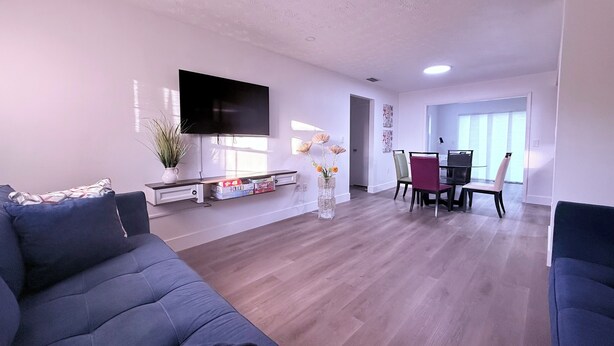 A modern living room with a flat-screen TV, a dining area with chairs, and a large window. - Sunny Oasis by Tarpon - Heated Pool, Hot Tub & Playground (Holiday)