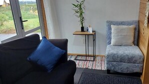 Une pièce avec un canapé noir, un fauteuil à motifs, une petite table sur laquelle est posée une plante, et une vue sur l’extérieur grâce à une grande fenêtre.