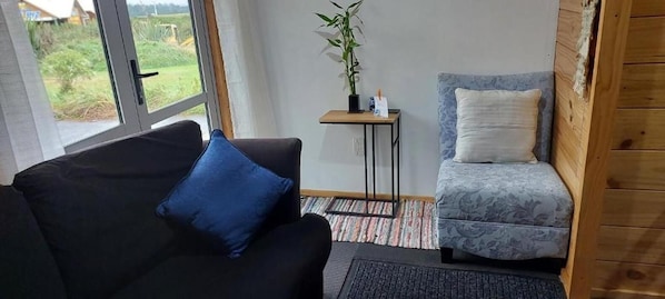 Une pièce avec un canapé noir, un fauteuil à motifs, une petite table sur laquelle est posée une plante, et une vue sur l’extérieur grâce à une grande fenêtre.