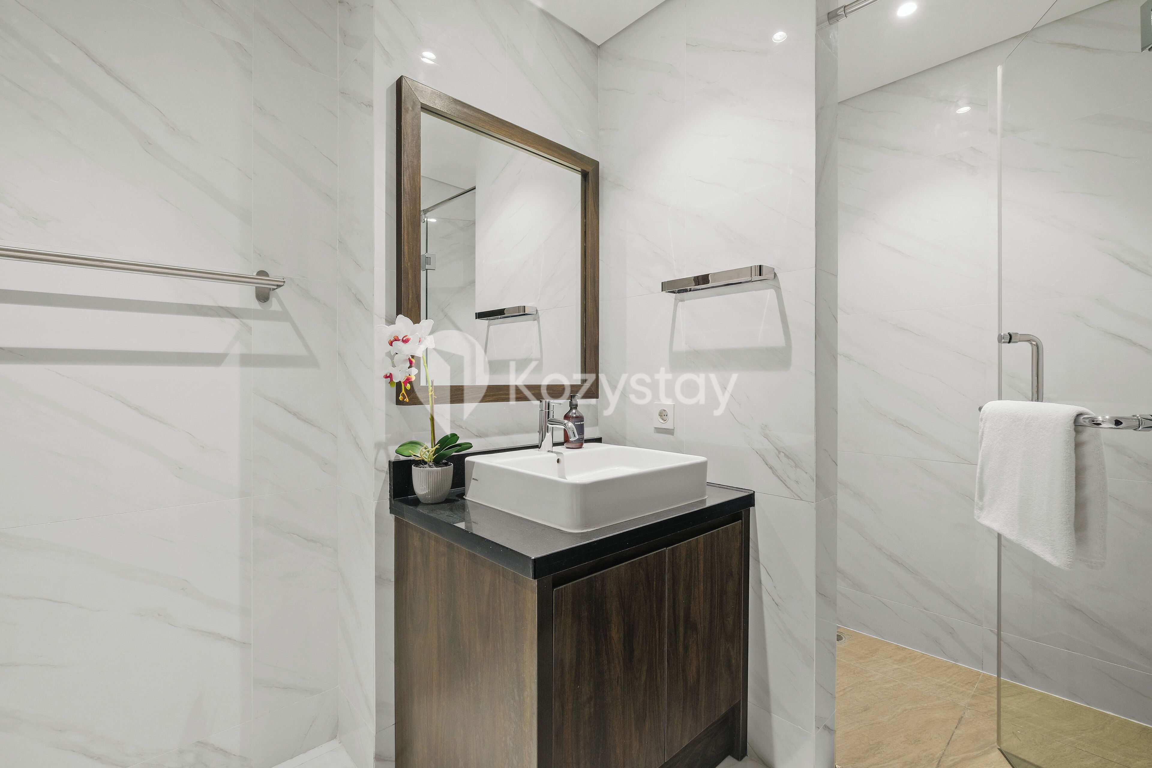 A modern bathroom with a marble wall, a wooden vanity, a white sink, a mirror, and a shower area.