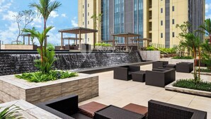 A rooftop patio with outdoor seating, a waterfall feature, and a modern building in the background. - Sorven by Kozystay - BSD (Pagedangan)