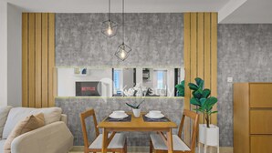 A modern living room with a wooden dining table, chairs, and a sofa. A plant is on the right side, and a TV is mounted on the wall. - Sorven by Kozystay - BSD (Pagedangan)