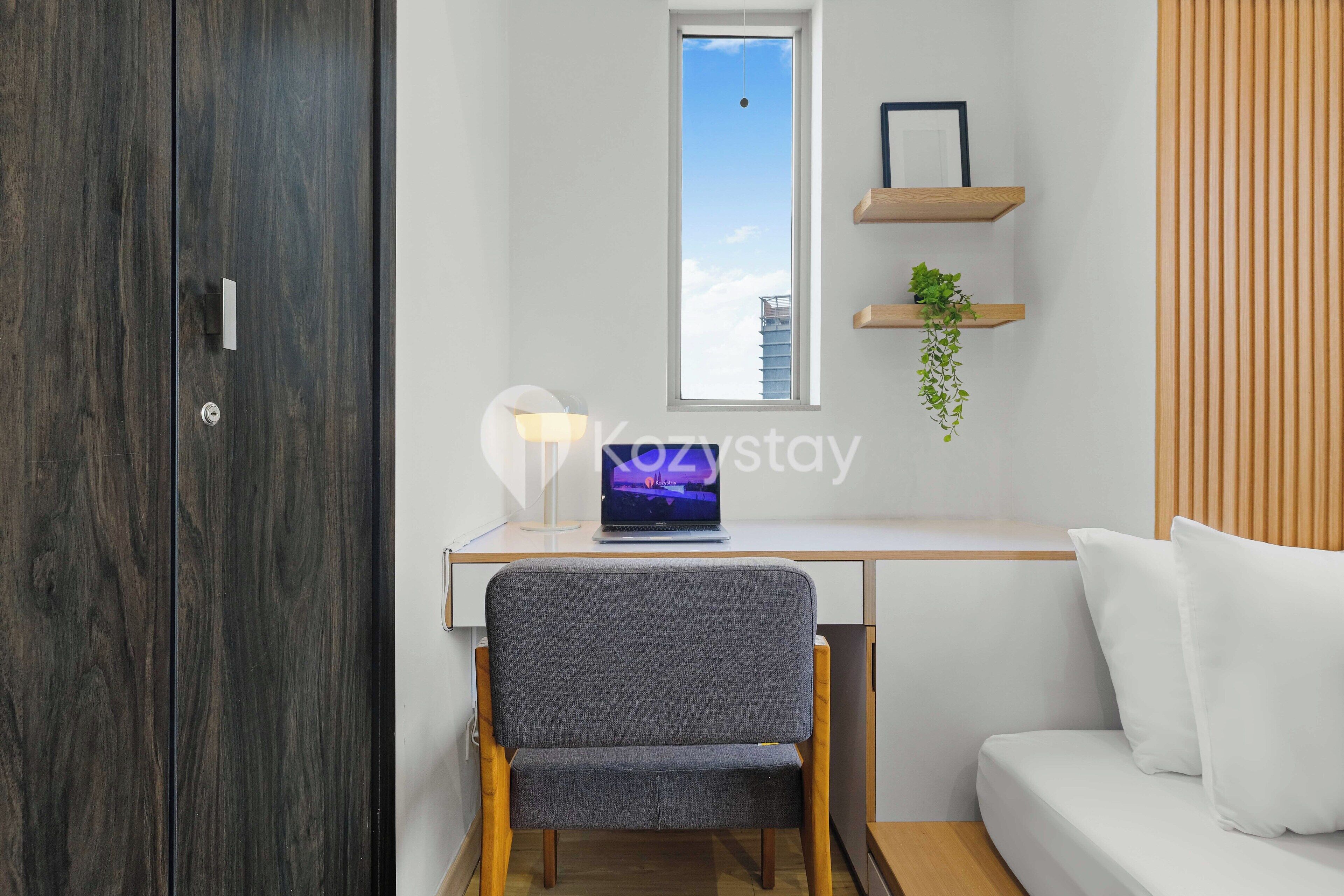 Une chambre moderne avec une armoire en bois, un bureau avec un ordinateur portable, une chaise, un canapé blanc et une fenêtre donnant sur le ciel.