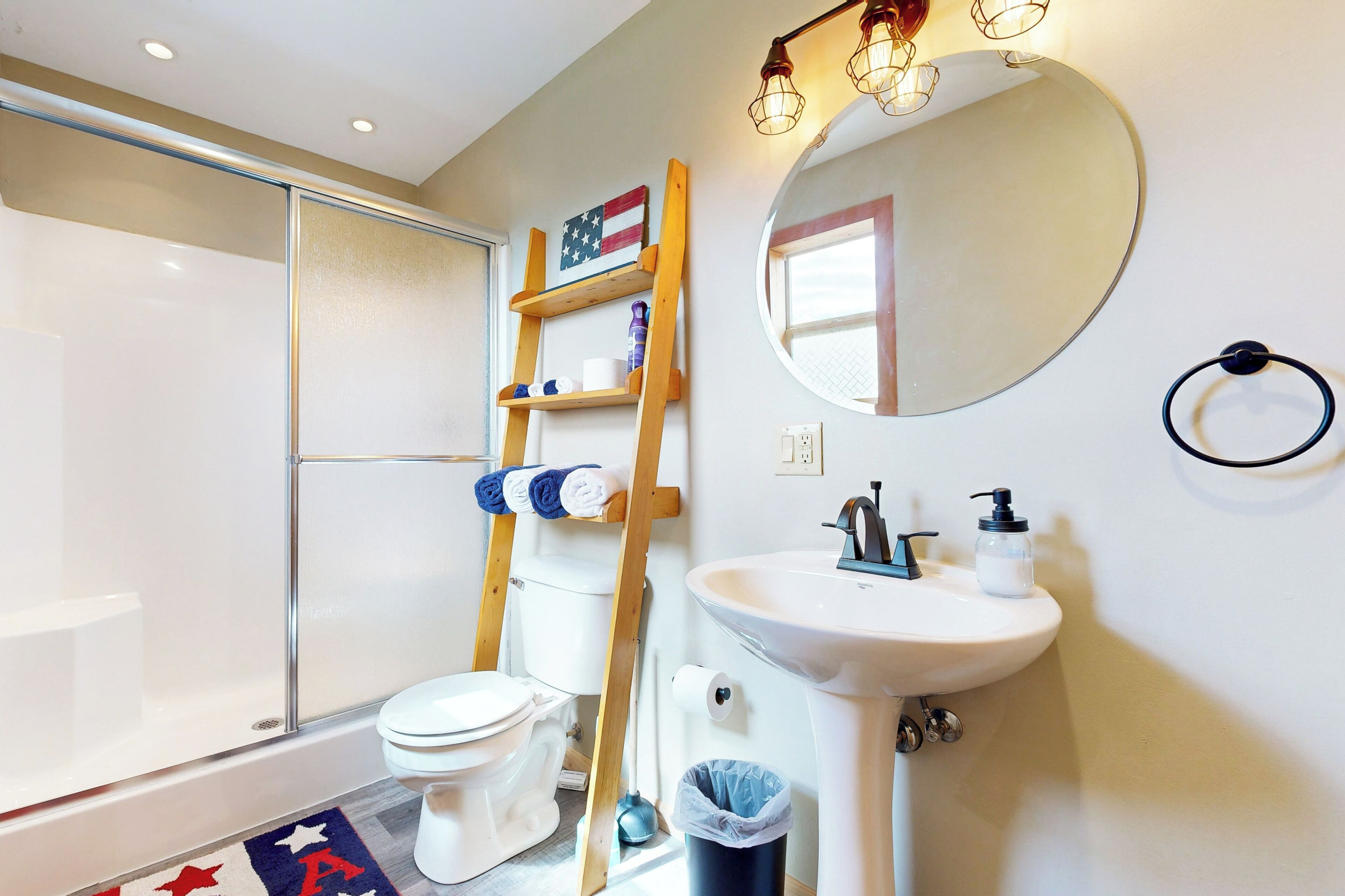 Un bagno con servizi igienici, lavandino e doccia. Si nota una scala di legno, un cestino e uno specchio a parete.