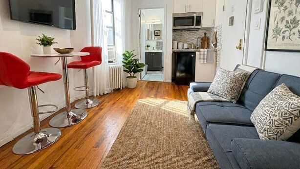 A modern living room with a grey sofa, red bar stools, a flat-screen TV, and a kitchen area with a microwave. - Sunny 1BR in Heart of Hell's Kitchen (New York)