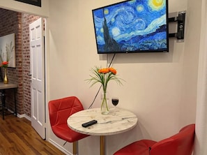 Ein Zimmer mit einem runden Tisch, roten Stühlen, einem Fernseher, der ein Gemälde zeigt, einer Vase mit Blumen und einem Weinglas.