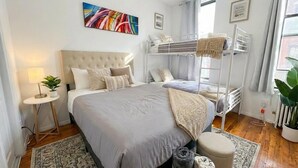 A bedroom with a large bed, a bunk bed, a bedside table with a lamp, a plant, and a painting on the wall. - Sunny 1BR in Heart of Hell's Kitchen (New York)