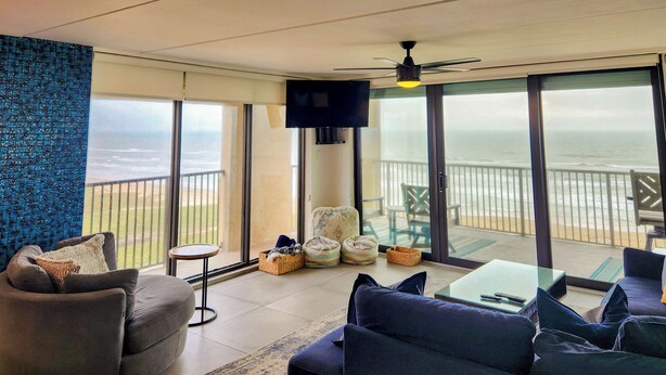 A living room with a sofa, armchair, and a glass table. There is a TV mounted on the wall and a balcony with a view of the ocean. - 3BR w/ Wraparound Balcony: Suntide III - Unit 901 (South Padre Island)