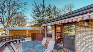 A wooden deck with a glass table and chairs, a brick building with a striped awning, and a view of trees and a distant building. - Work & Play! Spacious Lake-View Home in Greeley (Greeley)