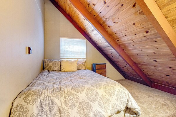 A bedroom with a sloped ceiling, a bed with patterned bedding, a window with blinds, and a small nightstand. - Dock! Dog-Friendly Lakefront Gem in Southern Maine (Waterboro)