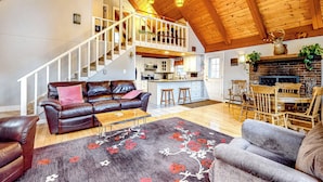 A spacious living area with a leather sofa, a grey armchair, and a wooden dining table with chairs. A staircase leads to an upper level. - Dock! Dog-Friendly Lakefront Gem in Southern Maine (Waterboro)