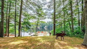 A forested area with a cabin and a boat docked by a lake. - Dock! Dog-Friendly Lakefront Gem in Southern Maine (Waterboro)