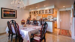 Un comedor con una araña, sillas de madera, una mesa con mantel floral y una cocina al fondo.