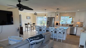 A modern kitchen with white cabinets, a built-in island, and stainless steel appliances. There is a dining area with white chairs and a table, and a living room with a grey sofa and a ceiling fan. - Gorgeous 3-bedroom pool, Direct gulf access In SE Cape Coral. Close to downtown (Cape Coral)