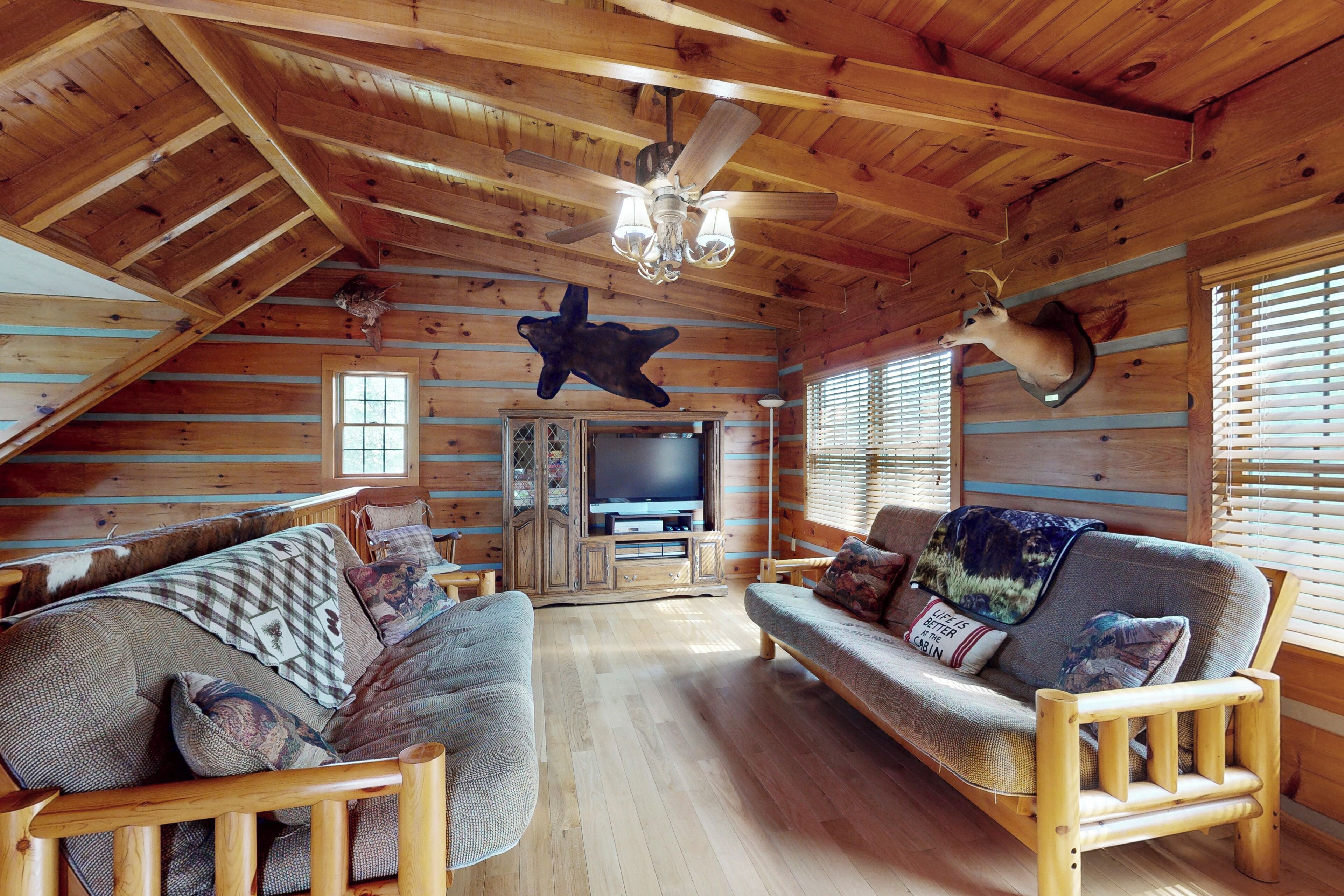 A log cabin interior with a wooden ceiling, a ceiling fan, a mounted television, and a sofa with decorative pillows.