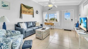 A living room with a blue sofa, a coffee table, and a TV. There is a ceiling fan, a window with a view, and a door. - Island Breeze Condo on the Beach (Englewood)