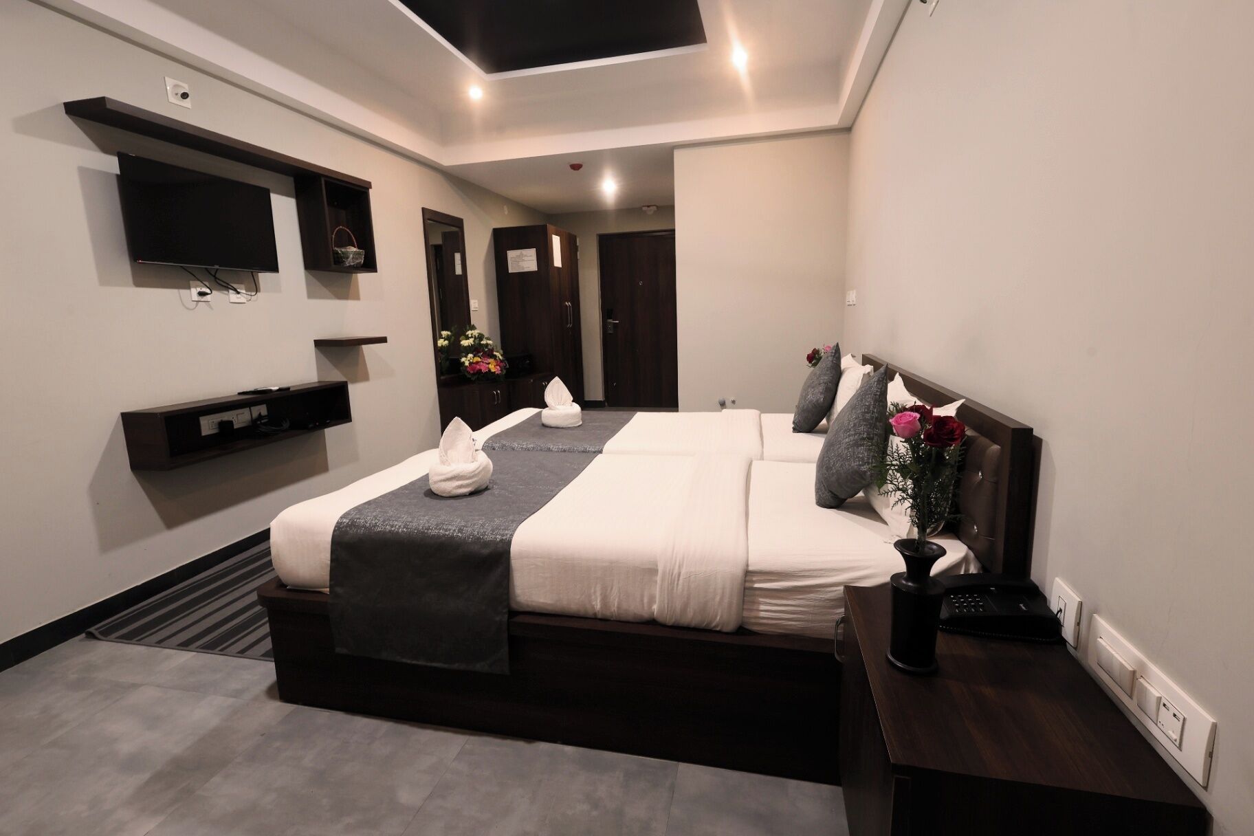 A modern hotel room with a large bed, a flat-screen TV mounted on the wall, a bedside table with a lamp, and a small desk with a computer.