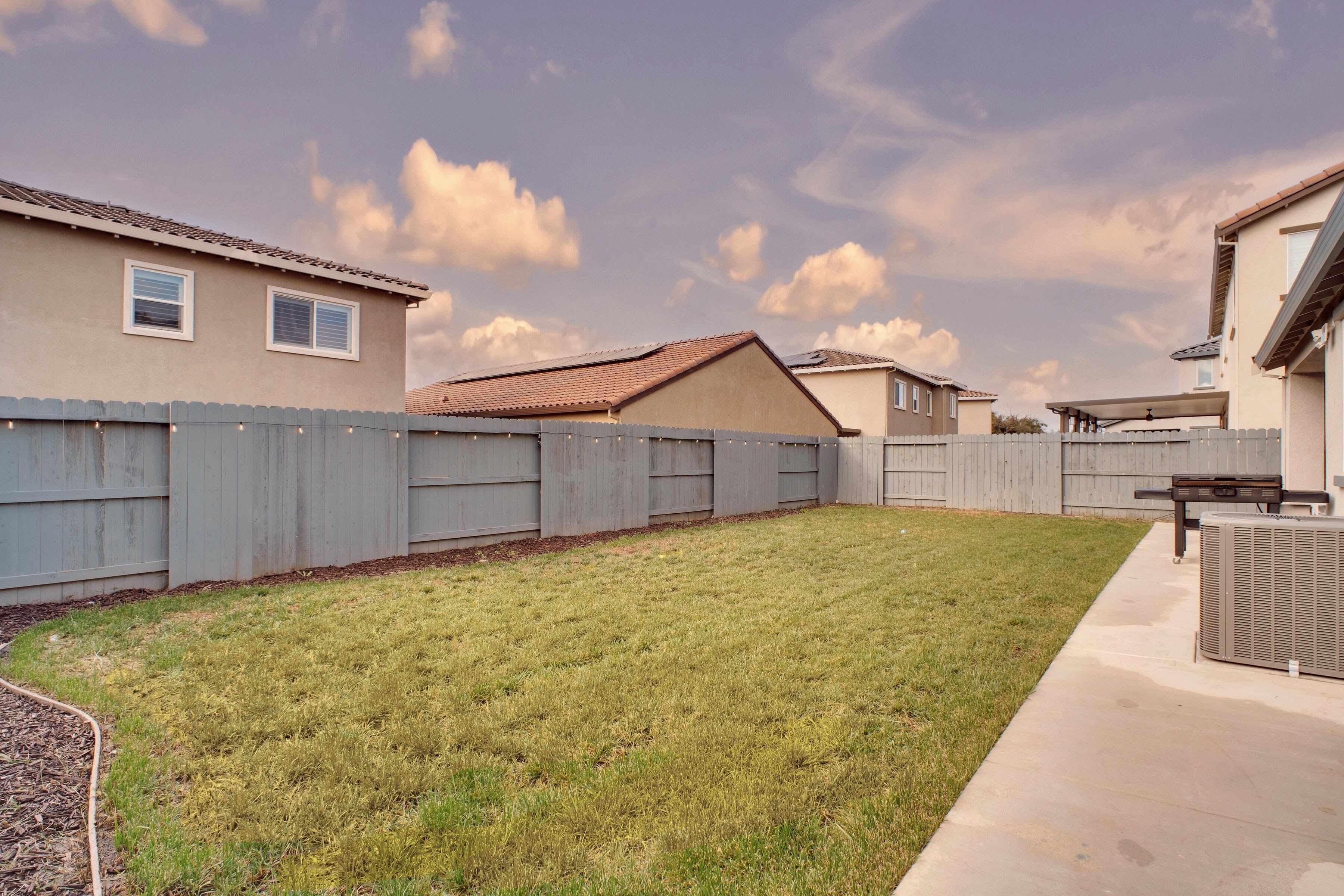 A residential backyard with a grassy lawn, a wooden fence, and a patio area with an air conditioning unit.