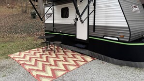 Un camper Coleman Datcher con veranda coperta, un'area pranzo con sedie e un tappeto a motivi su fondo di ghiaia.