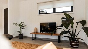 Sala de estar moderna com televisão de tela plana sobre um suporte de madeira, uma grande planta em vaso e uma janela com persianas.
