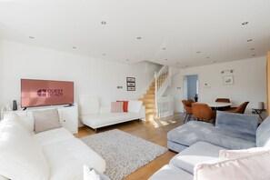 A modern living room with a flat-screen TV, white sofas, a staircase, and a dining area. - GuestReady - Riverside Retreat in Wapping (London)