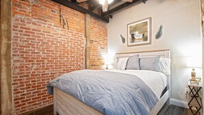 A bedroom with a brick wall, a bed with a blue and white bedspread, a wooden headboard, a nightstand with a lamp, and a framed picture on the wall.