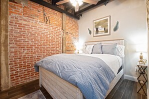 Une chambre avec un mur en briques, un lit recouvert d’une couvre-lit bleue et blanche, une tête de lit en bois, une table de chevet avec une lampe et un tableau encadré accroché au mur.