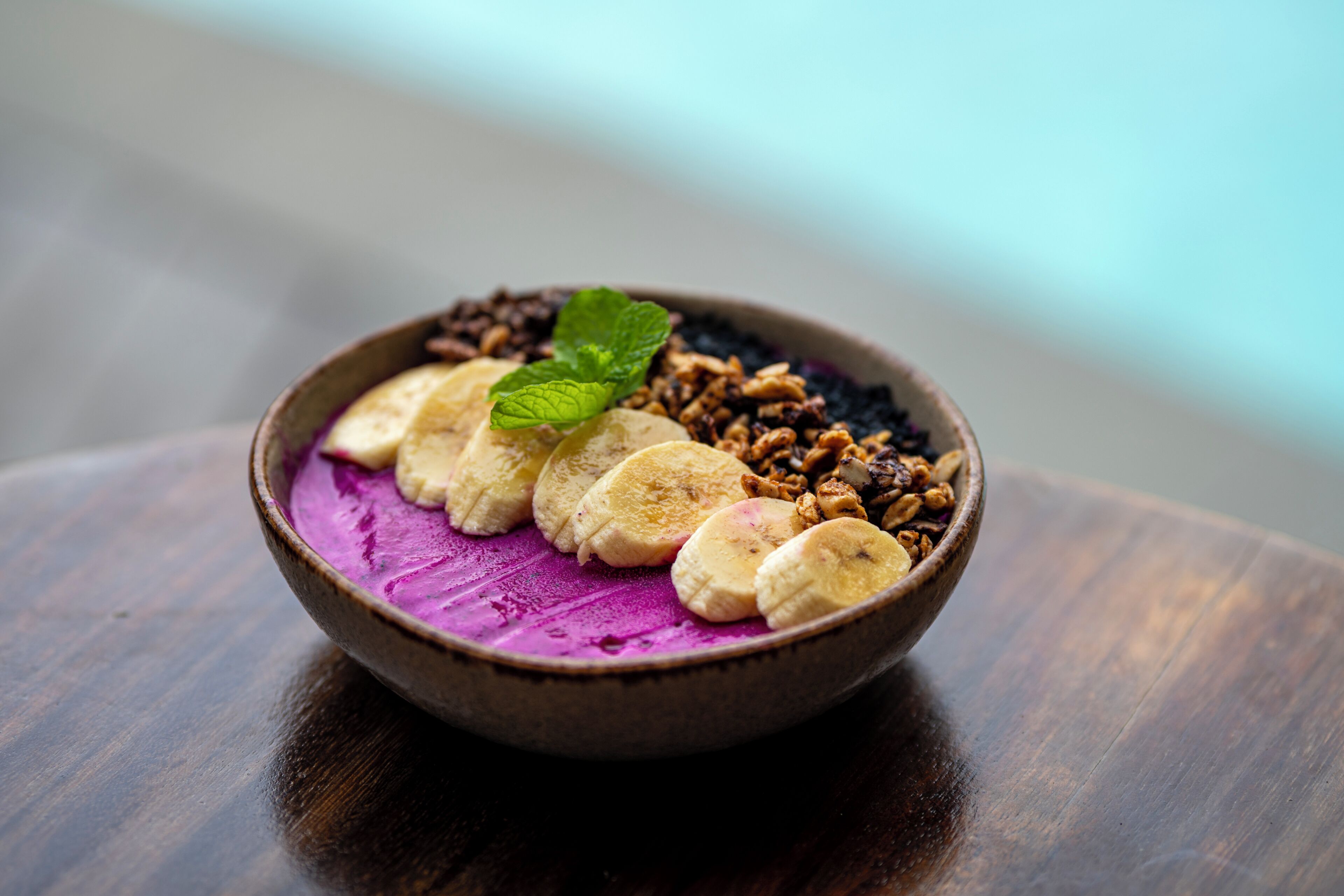Eine Schüssel mit Essen, Bananen, Granola und einer violetten Substanz auf einem Holztisch.