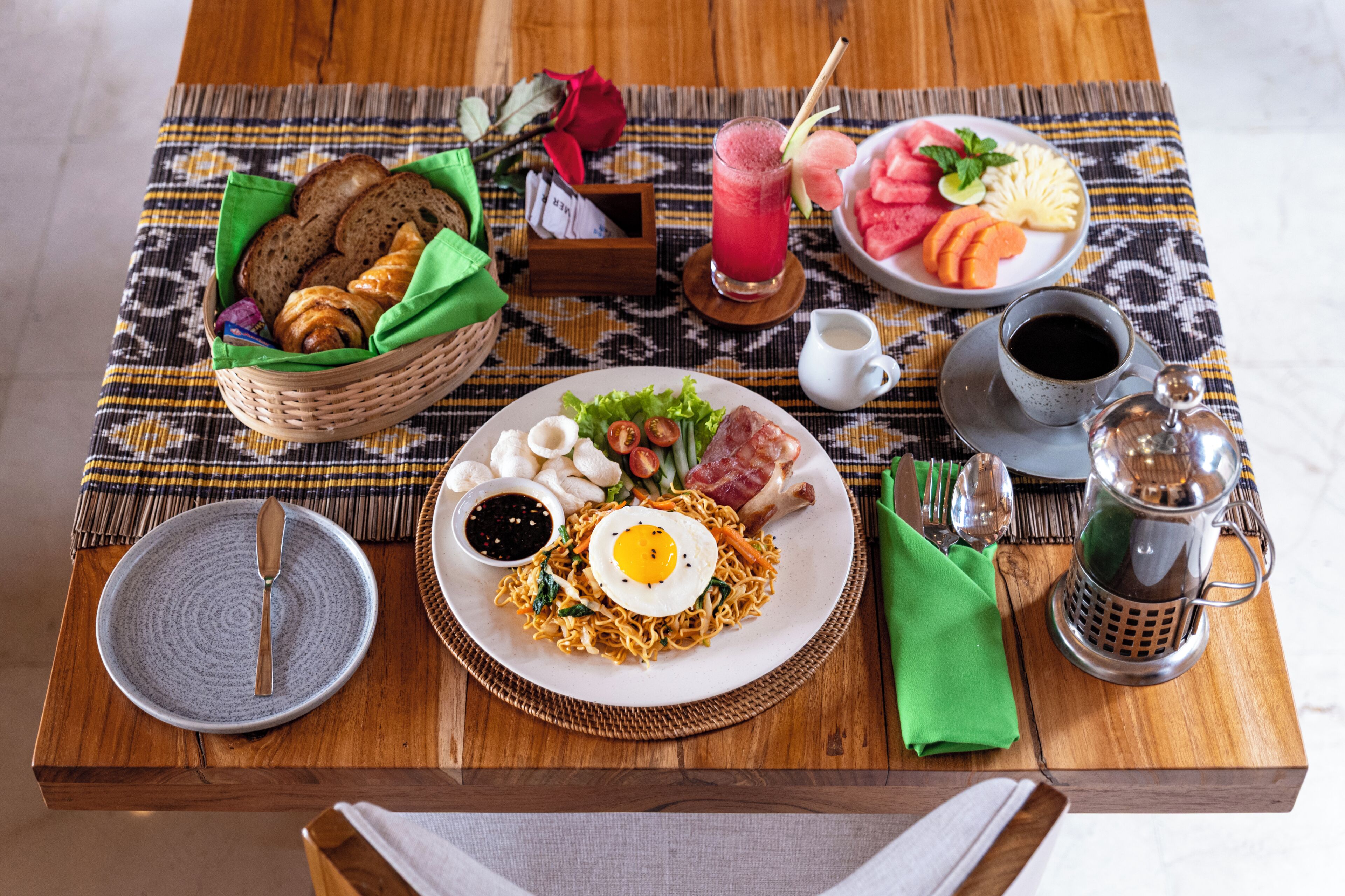 Una mesa de desayuno con diversos alimentos, incluyendo un plato de fideos con un huevo frito, un bol de fruta y un vaso de jugo.