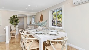 Espaço de refeições com uma mesa comprida posta para seis pessoas, rodeado por cadeiras de vime, e vista para o verde lá fora.