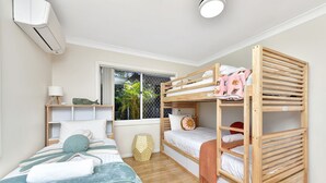 Quarto com beliche, uma cama de solteiro, mesa de cabeceira, luminária e janela com vista para área verde.