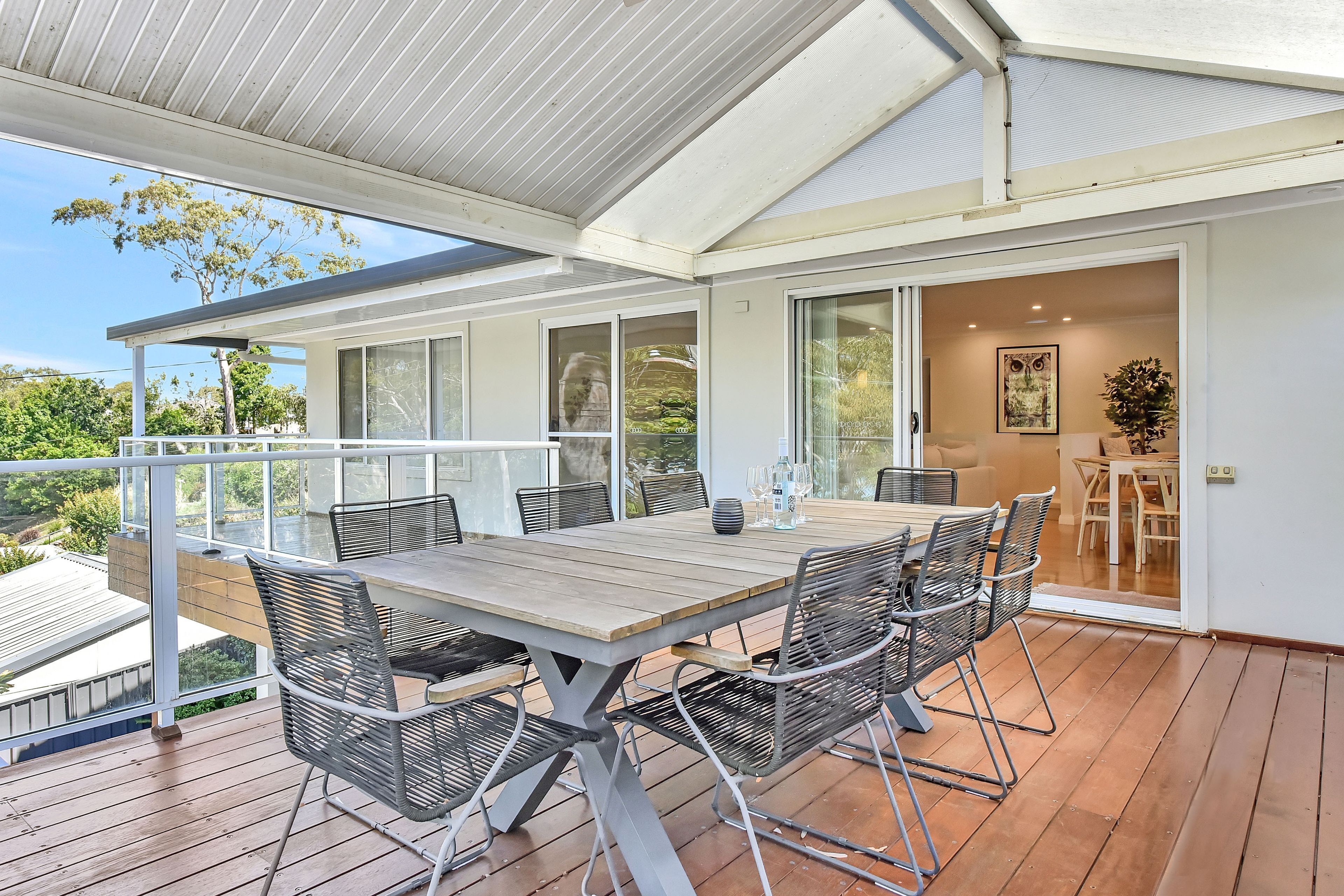 A covered outdoor dining area with a table and chairs, a glass balustrade, and a view of the surrounding greenery.