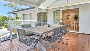 Uma área de refeições ao ar livre coberta, com mesa, cadeiras, guarda-corpo de vidro e vista para o verde ao redor.