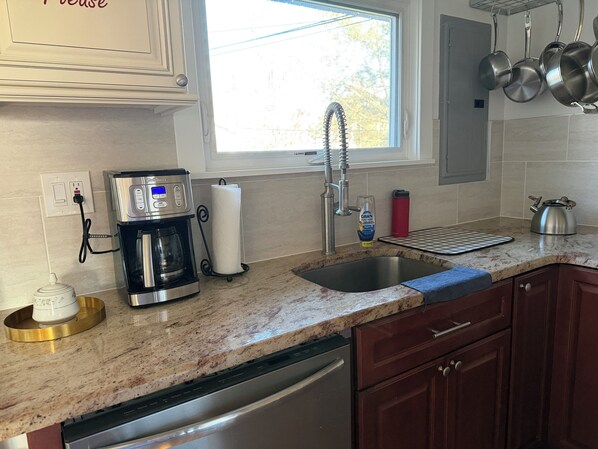 Una cocina con cafetera, lavabo, escurreplatos, hervidor y una botella de agua roja.