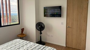 Ein Schlafzimmer mit Bett, Standventilator, montiertem Fernseher, Holztür und Fenster mit Blick auf Gebäude.