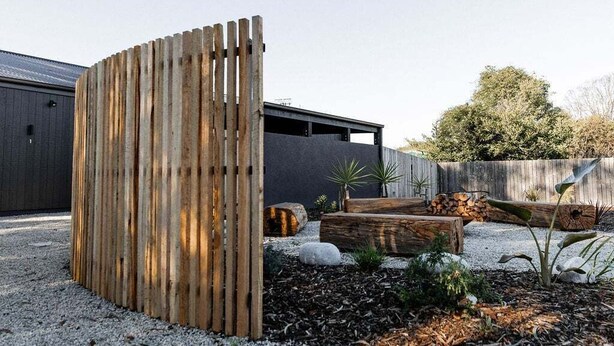 Een modern buitenverblijf met een houten omheining, een grindpad en een tuin met planten en boomstammen.