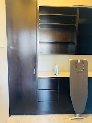 Ein kompakter, moderner Raum mit einem dunkelbraunen Kleiderschrank, einem kleinen Bügelbrett und einer weißen Arbeitsplatte.
