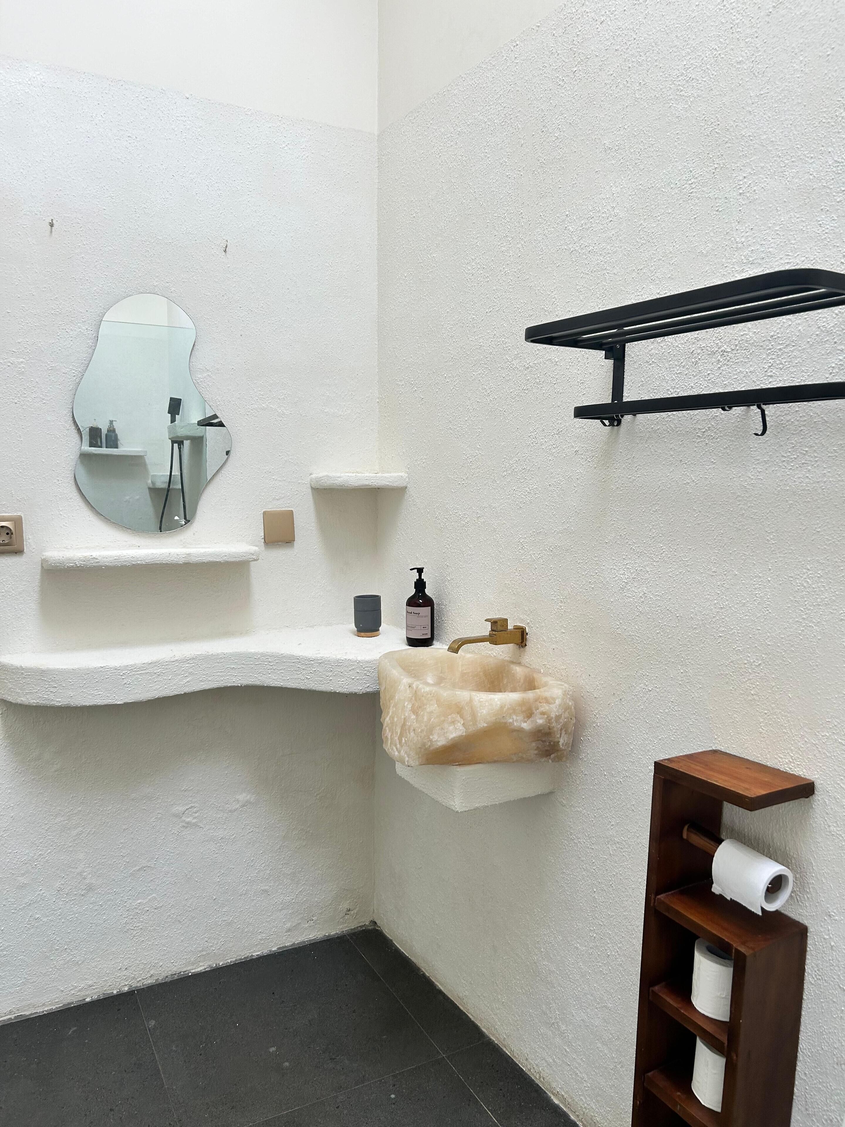 A bathroom with a white textured wall, a sink with a mirror, a soap dispenser, and a shelf with a towel.