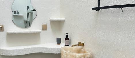 A bathroom with a white textured wall, a sink with a mirror, a soap dispenser, and a shelf with a towel.
