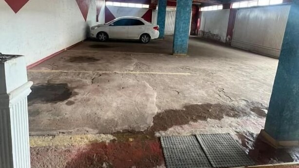 An indoor parking area with a white car parked in the center. The floor is concrete with visible stains and patches. There are blue pillars supporting the ceiling, and the walls are painted in red and white. - Caribe Palace Hotel Samambaia Sul (Samambaia)