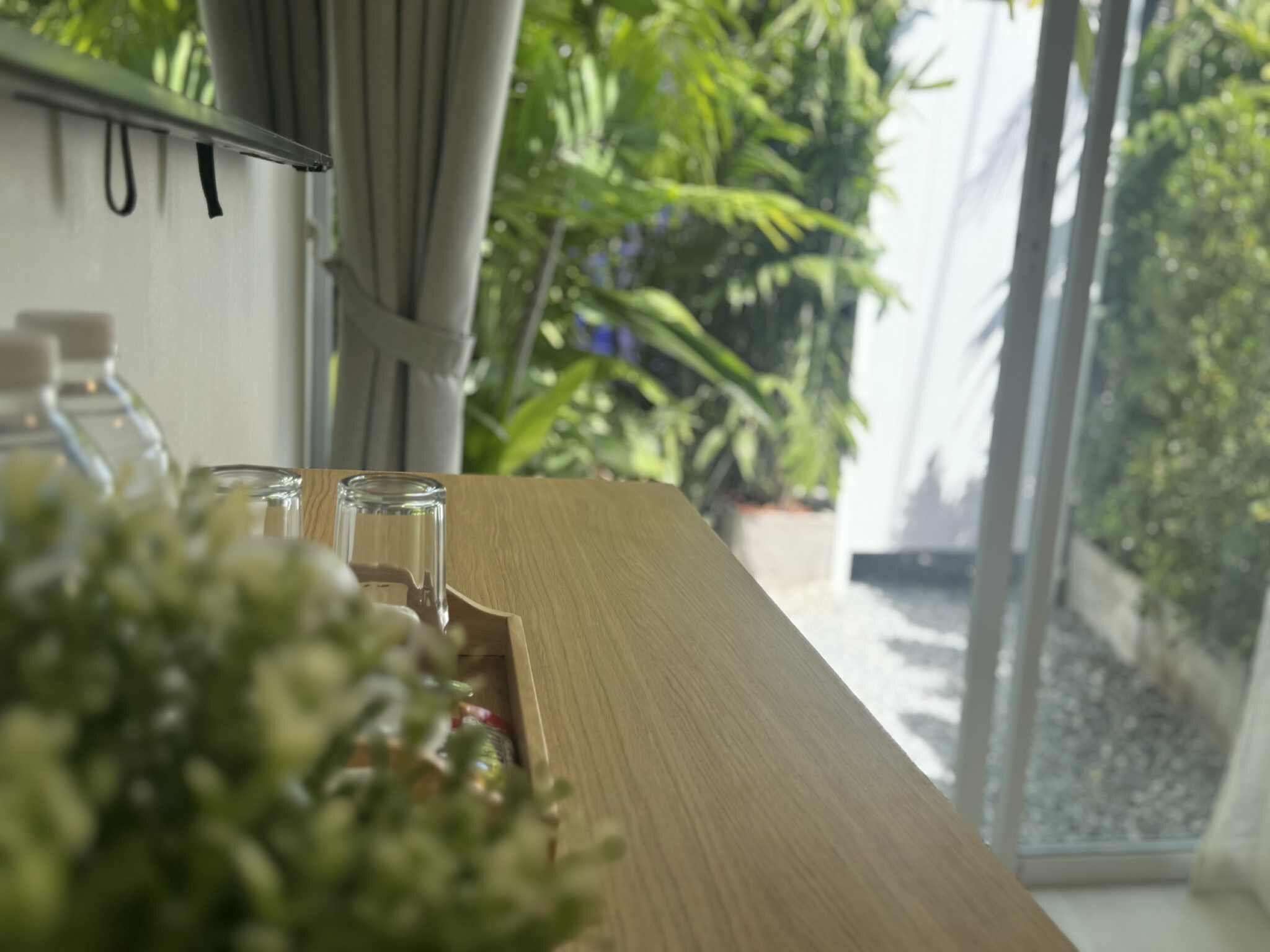 A wooden table with a glass container and a bottle, a window with curtains, and a view of greenery outside.
