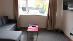 Huoneessa on harmaa sohva, värikäs pöytä, ikkuna, jossa on verhot, ja seinällä oleva televisio.