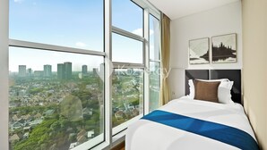 Ein Schlafzimmer mit großem Fenster und Blick auf die Stadt, einem ordentlich bezogenen Bett sowie zwei an der Wand befestigten Kunstwerken.