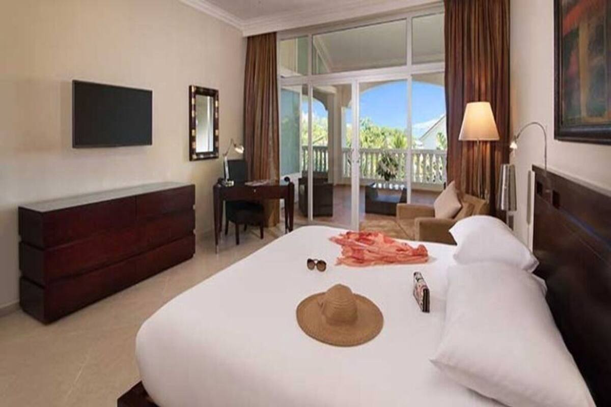 Ein Hotelzimmer mit einem großen Bett, einem Schreibtisch, einem Sessel, einem Fernseher und einem Balkon mit Blick auf Grünflächen.